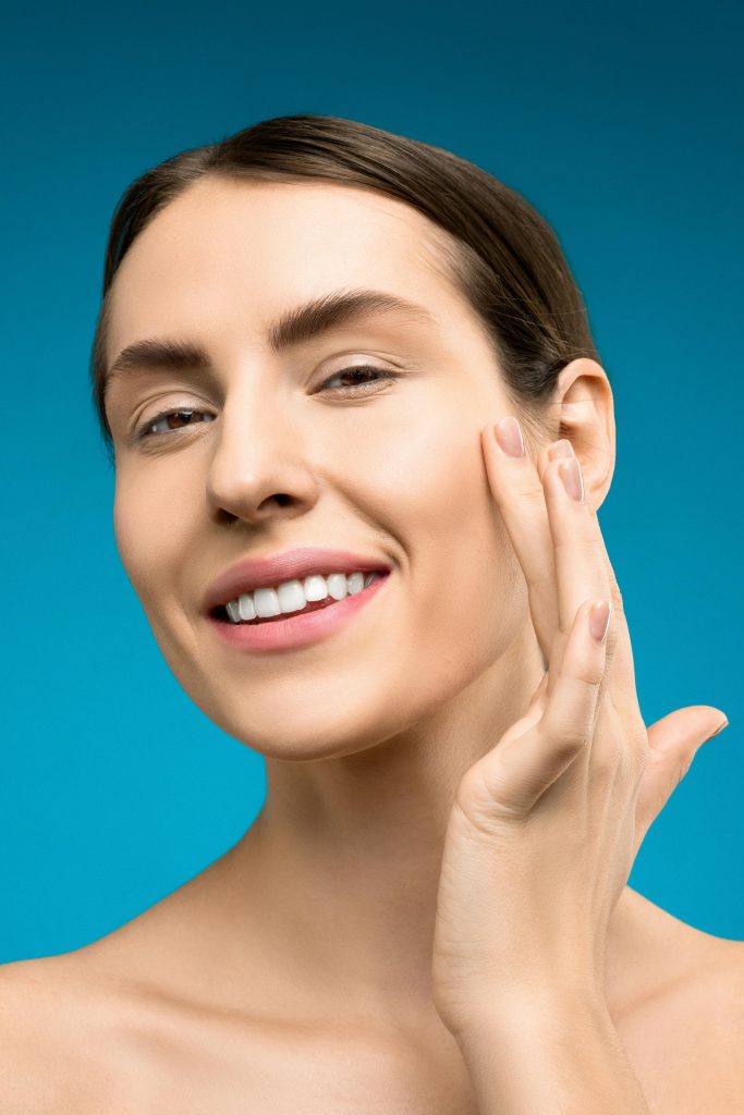 pexels-photo-3762185-3762185 Close-up of a young woman with smooth skin smiling brightly against a blue background.