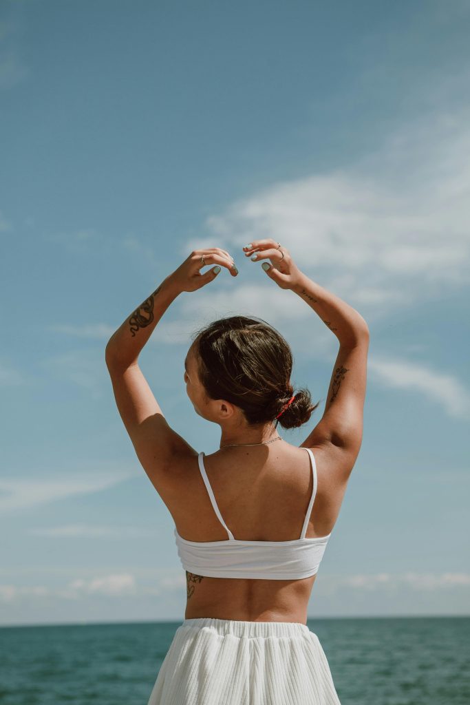 pexels-photo-4960254-4960254 A woman raises her arms freely under the bright summer sky by the ocean.